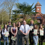 A group of approximately seven students and the college Principal Marcus Clinton standing outside on a lawn in front of a large, historic red-brick building. Students are holding white certificates and smiling for a group portrait.