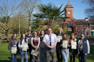 A group of approximately seven students and the college Principal Marcus Clinton standing outside on a lawn in front of a large, historic red-brick building. Students are holding white certificates and smiling for a group portrait.