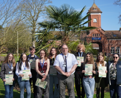A group of approximately seven students and the college Principal Marcus Clinton standing outside on a lawn in front of a large, historic red-brick building. Students are holding white certificates and smiling for a group portrait.