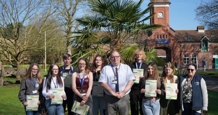 A group of approximately seven students and the college Principal Marcus Clinton standing outside on a lawn in front of a large, historic red-brick building. Students are holding white certificates and smiling for a group portrait.